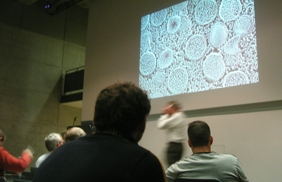 An image of people sitting in a lecture hall, with an enlarged photo of cells projected against the wall.