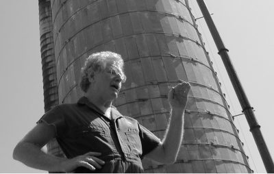 A black and white picture of a person wearing overalls standing in front of an agricultural silo.