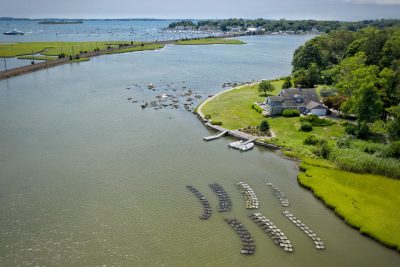 An arial image of a fishery in green water located next to a coastal home with a bay of ships in the background.